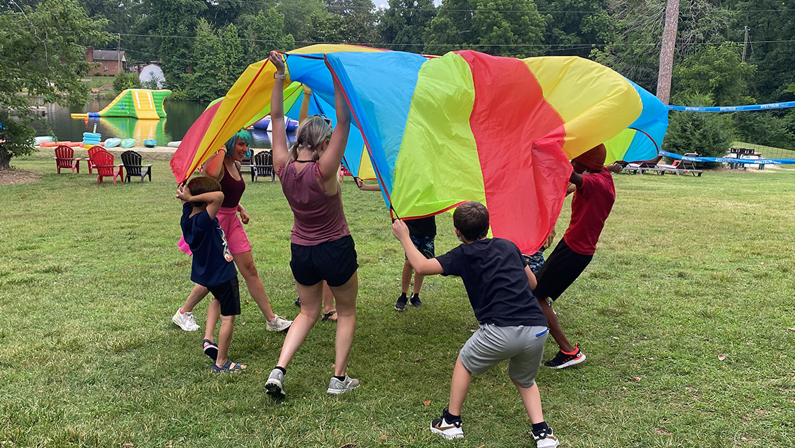 Children and CSD masters students play outside with a rainbow-colored parachute